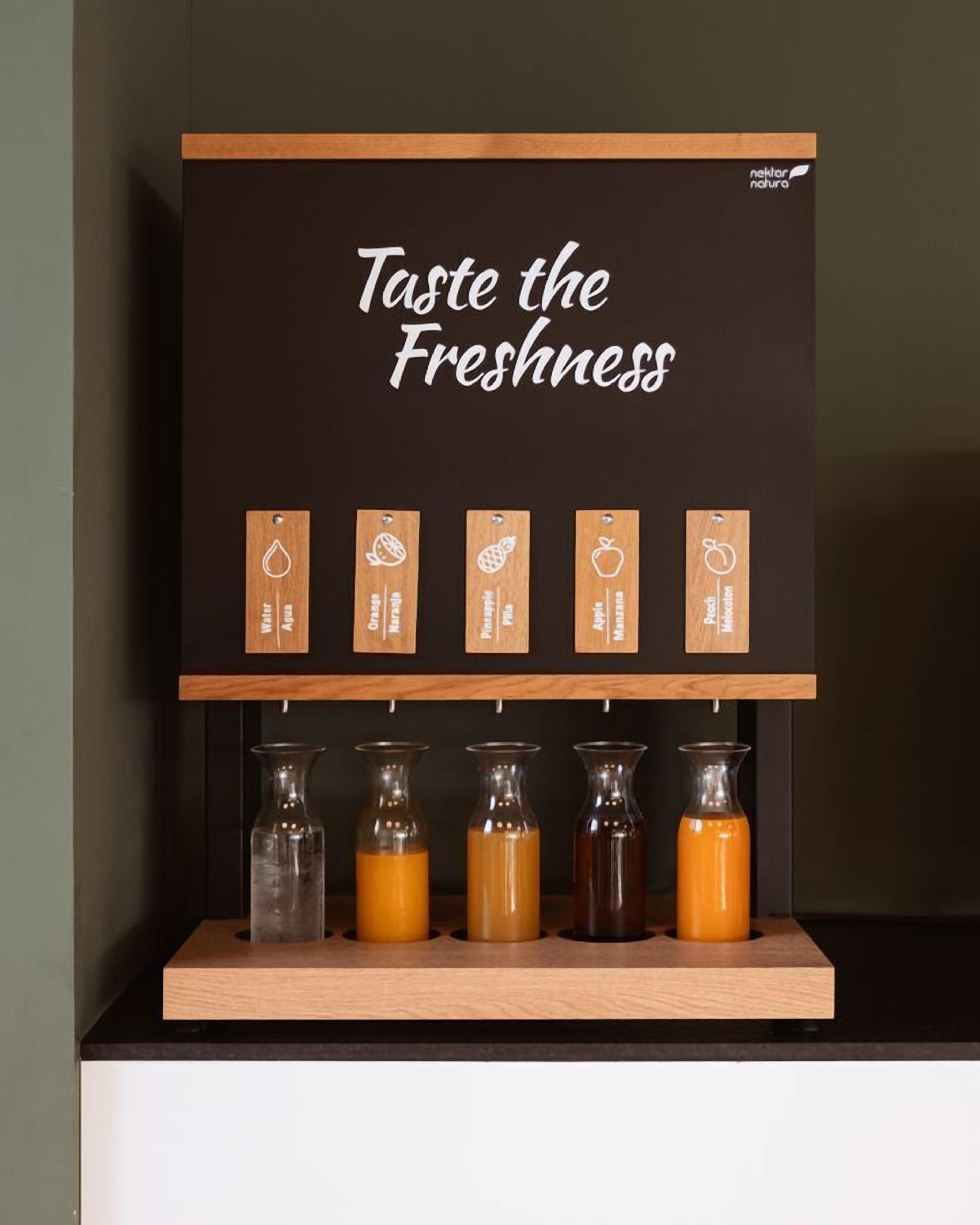 Un expositor con el texto “Saborea la Frescura” y etiquetas de madera con nombres de frutas como Melón, Naranja, Piña, Manzana y Melocotón. Debajo, hay cinco botellas de vidrio con líquidos de diferentes colores.