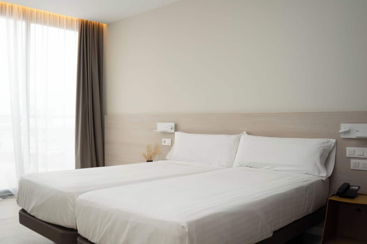Een hotelkamer met een tweepersoonsbed, een nachtkastje met lamp, een telefoon aan de muur en een raam met dunne gordijnen.