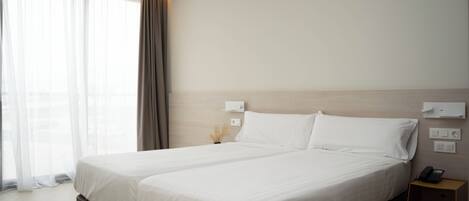 Habitación de hotel con cama doble, mesita de noche con lámpara, teléfono en la pared y ventana con cortinas translúcidas.