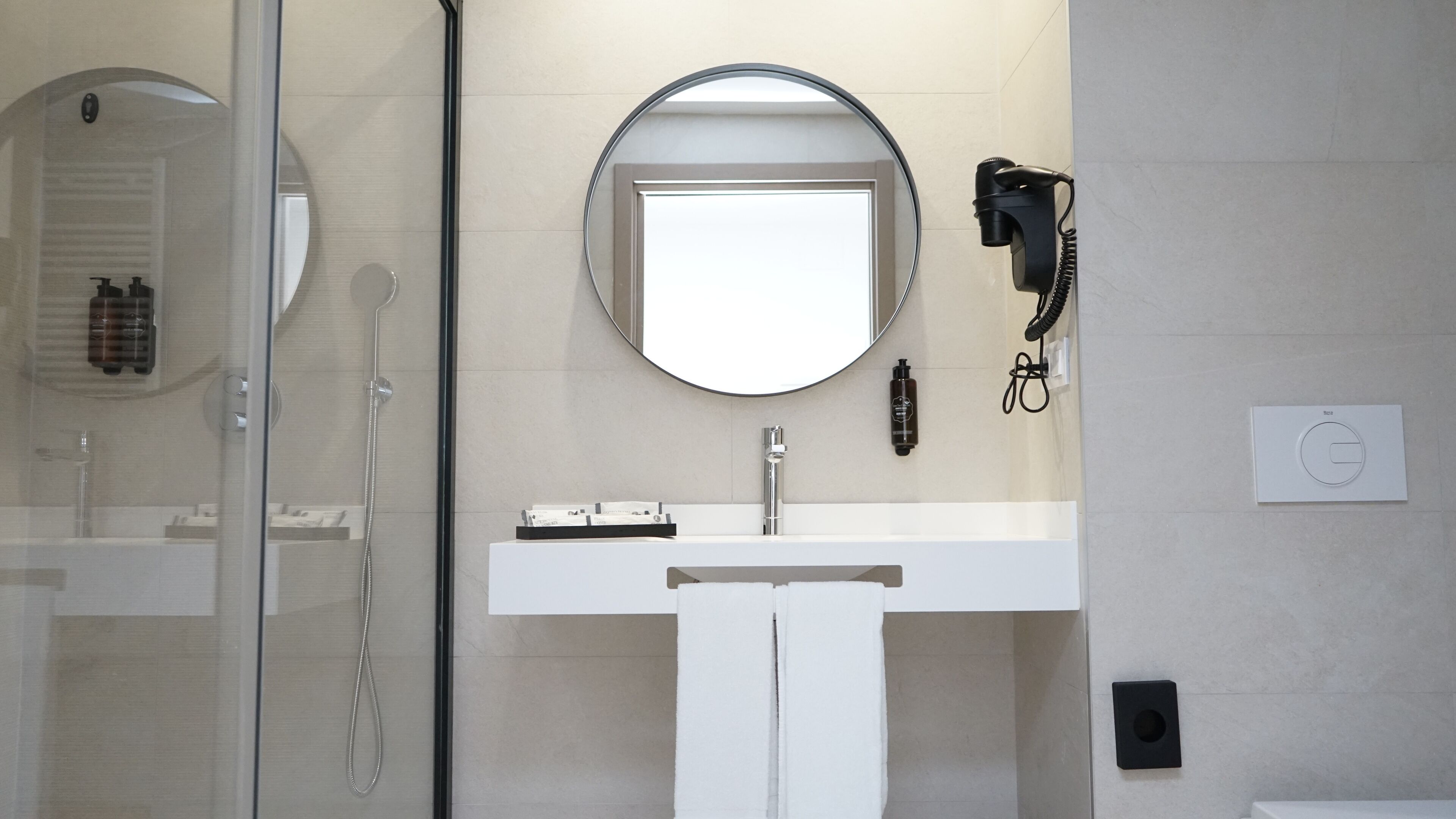 A modern bathroom with a glass shower enclosure, a white sink, a round mirror, and a hairdryer on the wall.