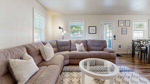 A living room with a large L-shaped sectional sofa, a glass coffee table, and a dining area with a table and chairs. - Modern Beach Cottage with a covered patio fire pit grill and easy beach access (Rockaway Beach)