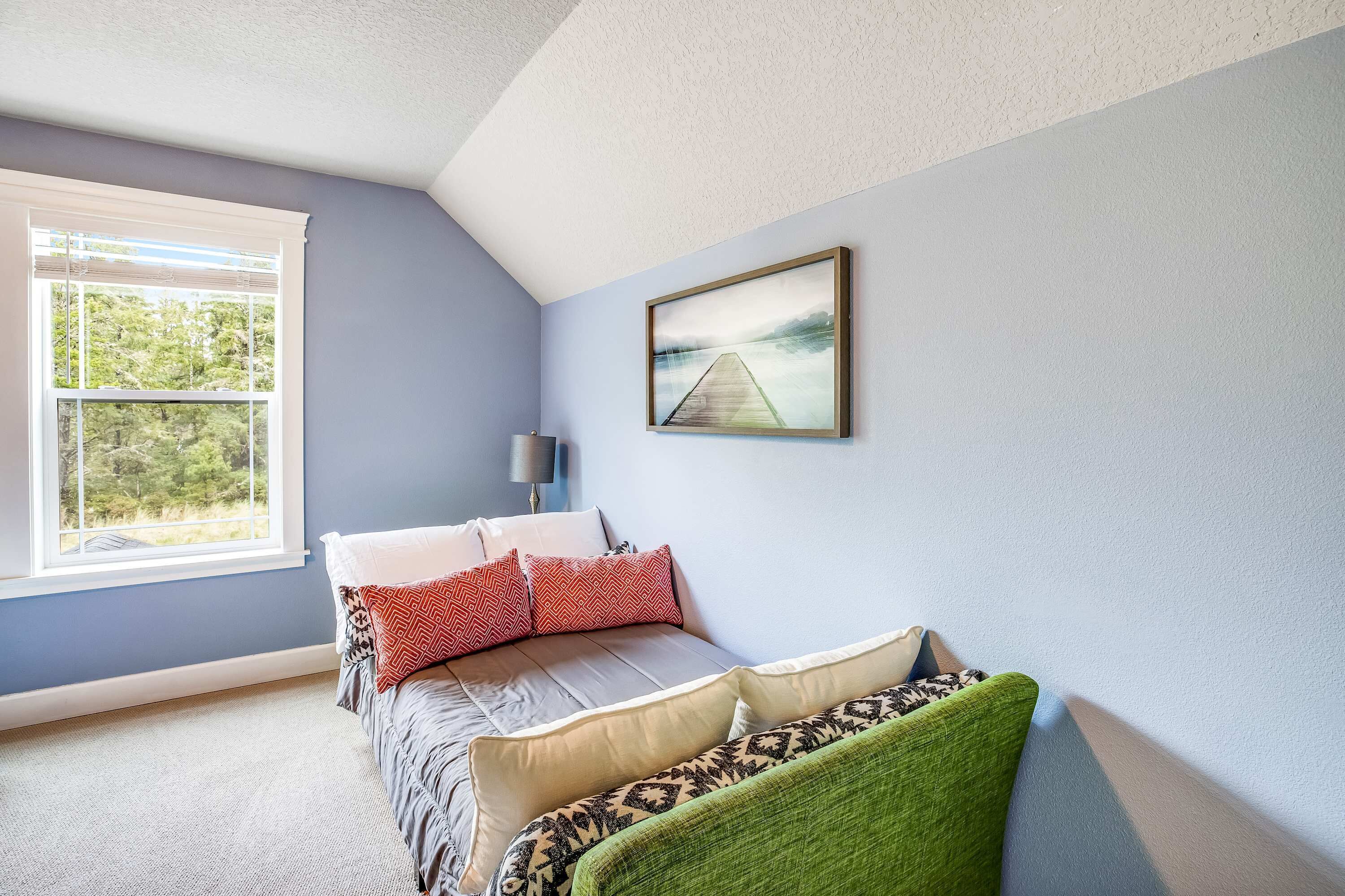 A bedroom with a bed, pillows, a green chair, a painting, and a window with a view of trees.