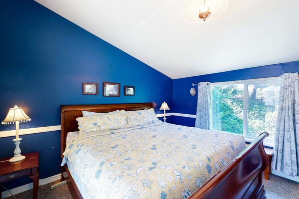 A bedroom with a bed, two bedside lamps, a window with curtains, and a view of greenery outside.