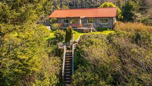 A house with a red roof, surrounded by dense greenery and a wooden staircase leading to a deck. - Delightful dog friendly home with bay views private hot tub and sauna (Rockaway Beach)