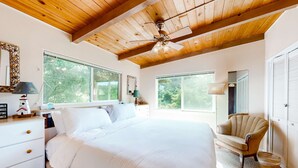 Ein Schlafzimmer mit einem großen Bett, Holzbalkendecke und Deckenventilator. Zwei Fenster sorgen für natürliches Licht und einen Blick ins Grüne.