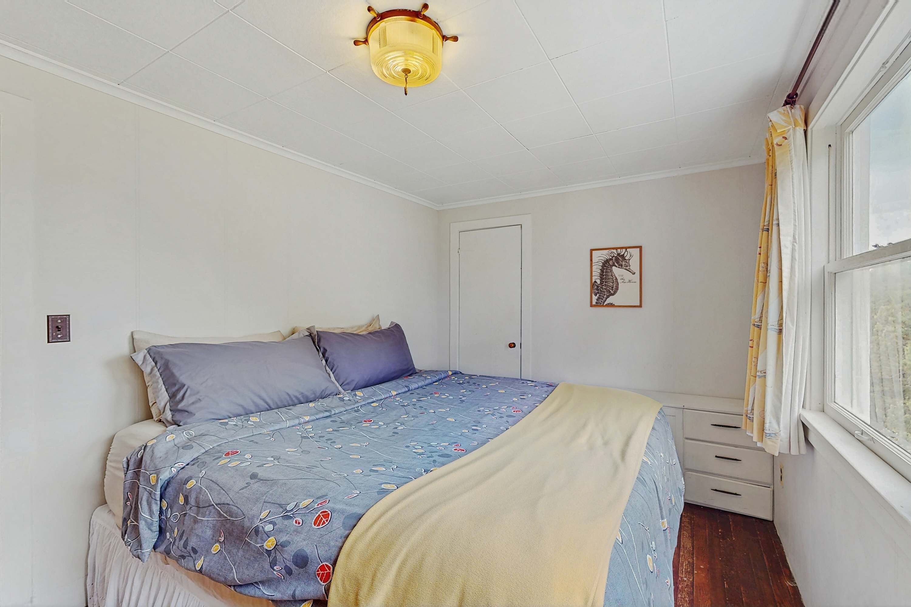 Een slaapkamer met een bed, nachtkastje, raam met gordijnen, een deur en een plafondlamp.