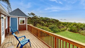 A wooden deck with blue chairs, a small building with a blue door, and a view of the ocean and greenery.
