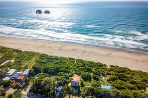 An aerial view of a beach with a dense forest area in the foreground, featuring several buildings with flat roofs. - McKinnon's beach house oceanfront with beach views deck patio and fireplace dog friendly (Rockaway Beach)