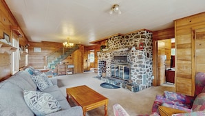 A cozy log cabin interior with a stone fireplace, wooden furniture, and a staircase leading to an upper level.