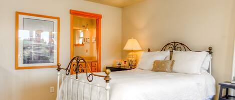 A bedroom with a white bed, a nightstand with a lamp, a framed picture on the wall, and a door with an orange frame.