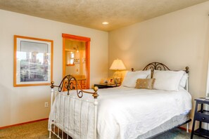 A bedroom with a white bed, a nightstand with a lamp, a framed picture on the wall, and a door with an orange frame. - Beachfront home with private beach access fireplace washer dryer yard deck (Rockaway Beach)