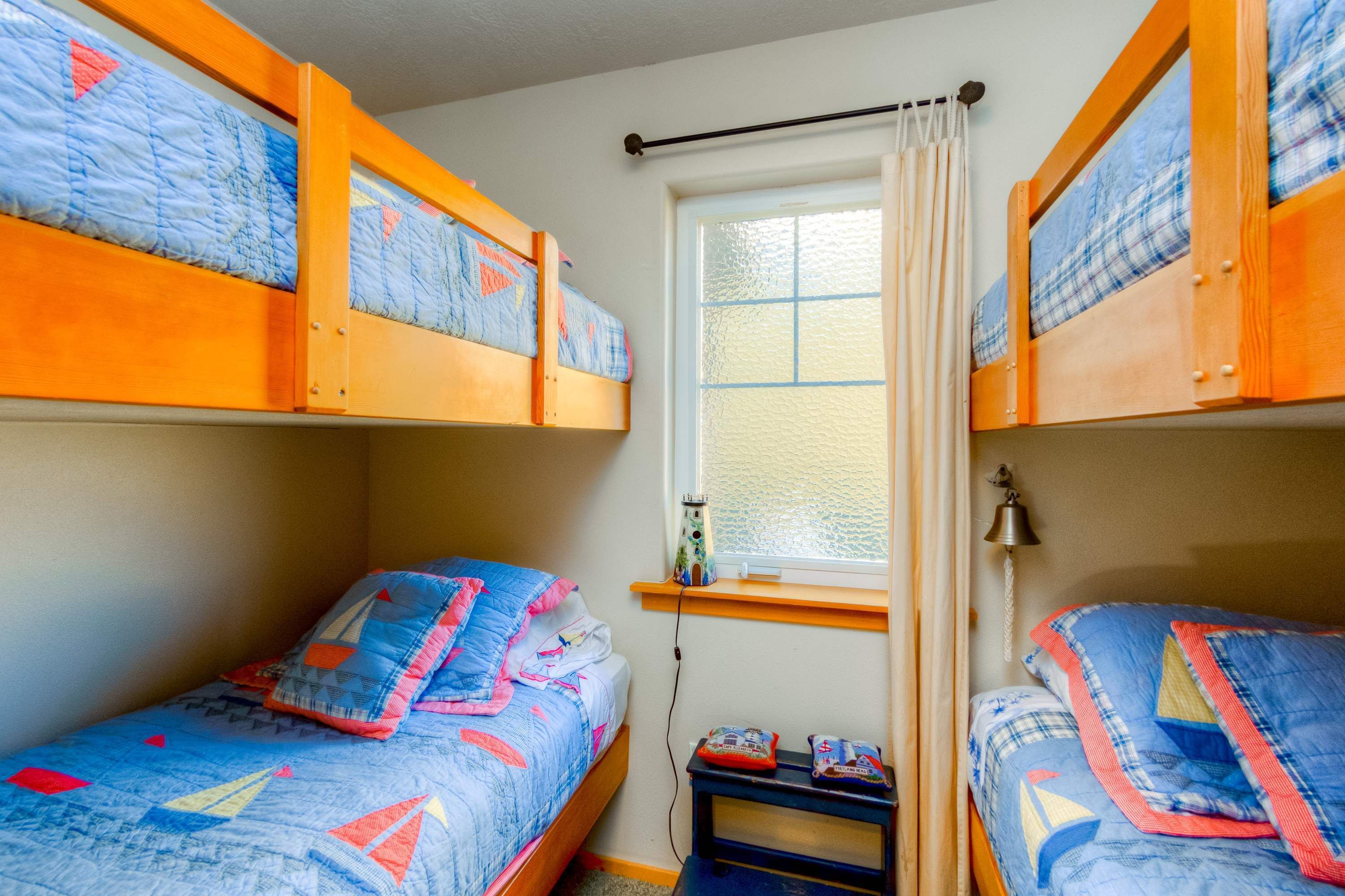Une chambre avec deux lits, chacun recouvert d’une courtepointe bleue ornée de motifs de voiliers. Il y a une fenêtre avec un rideau, une petite table avec une lampe et une étagère au-dessus de la fenêtre.