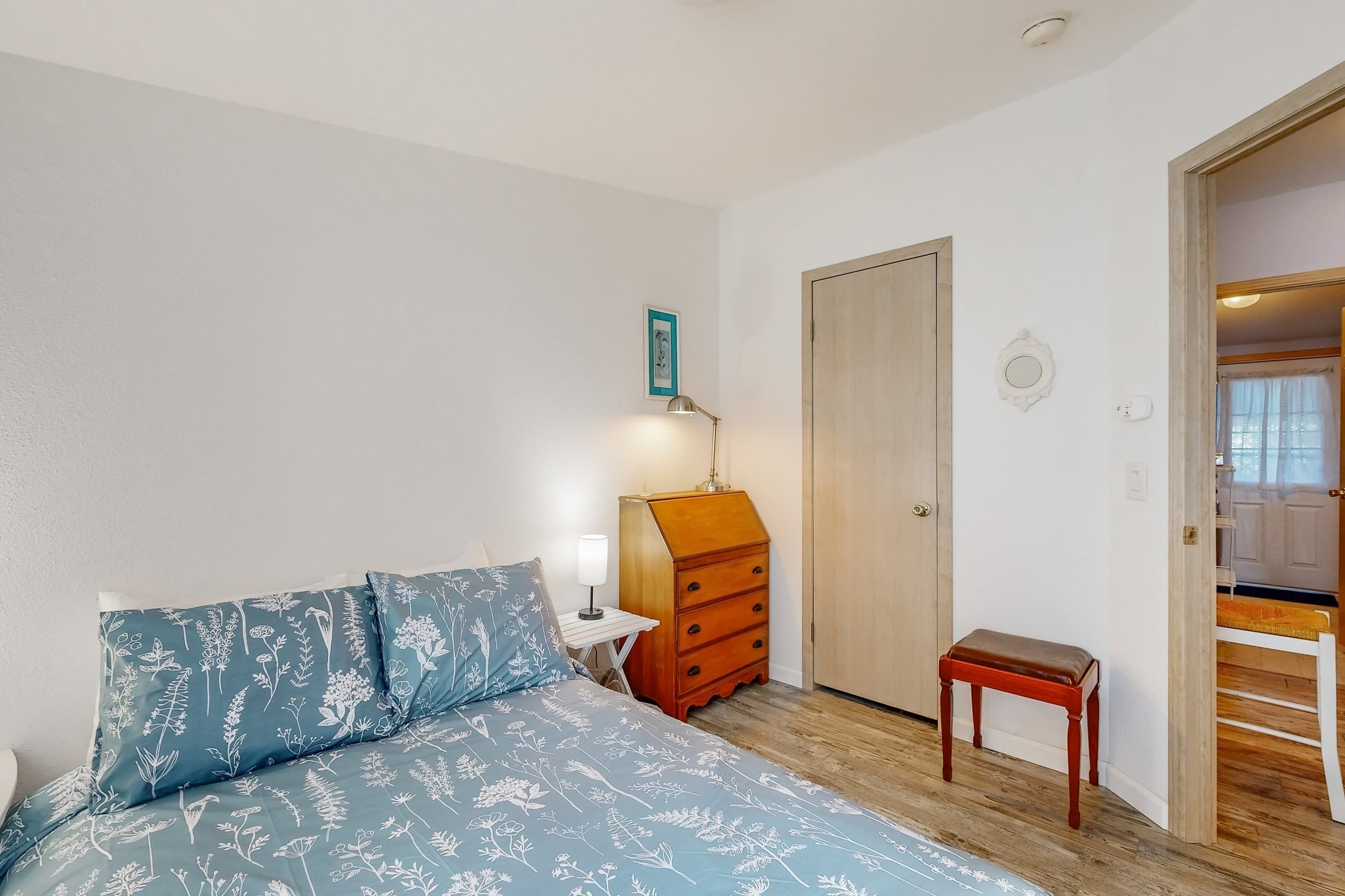 Une chambre à coucher comprenant un lit, une commode, une table de chevet, une lampe et une porte donnant sur une autre pièce.