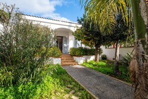 A white building with a covered entrance, surrounded by lush greenery and a gravel path leading to the door. - Holiday Home 'Villa Cala Azzurra Porto Cesareo' with Private Garden and Air Conditioning (Scala di Furno)