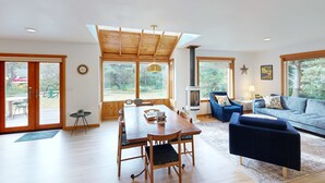 A spacious living area with a wooden dining table, blue sofas, and a large window offering a view of the outdoors. - Dog friendly home close to the beach with patio BBQ and WiFi (Rockaway Beach)