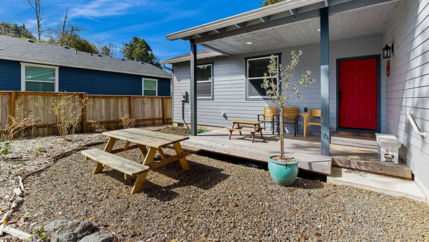 Ein Haus mit überdachter Veranda, einer roten Tür und einem kleinen Garten mit Holztisch und -stühlen.