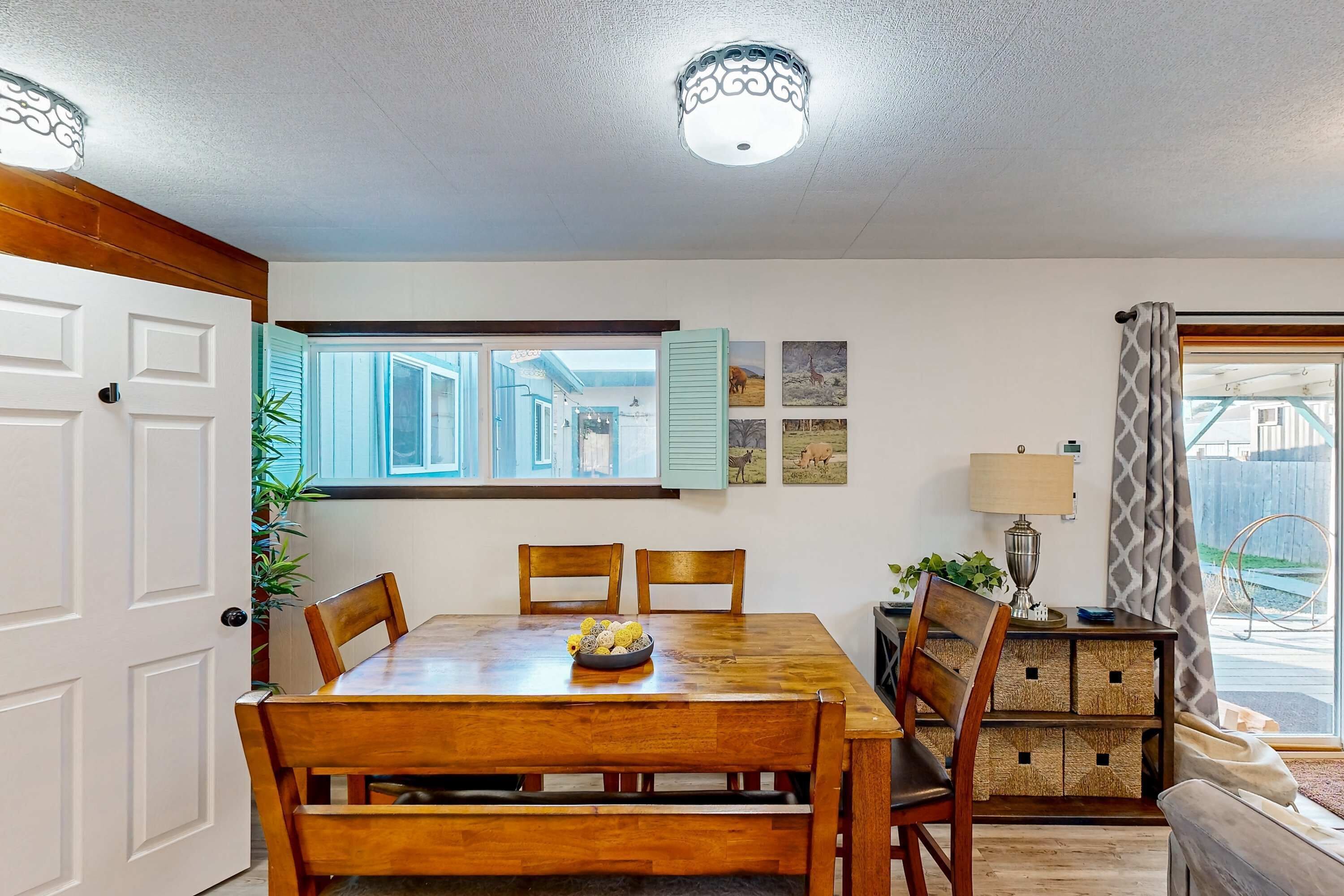 A dining area with a wooden table, chairs, and a bowl of fruit. There is a window with shutters, a lamp, and a plant.