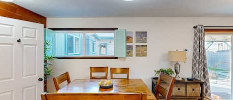 A dining area with a wooden table, chairs, and a bowl of fruit. There is a window with shutters, a lamp, and a plant.