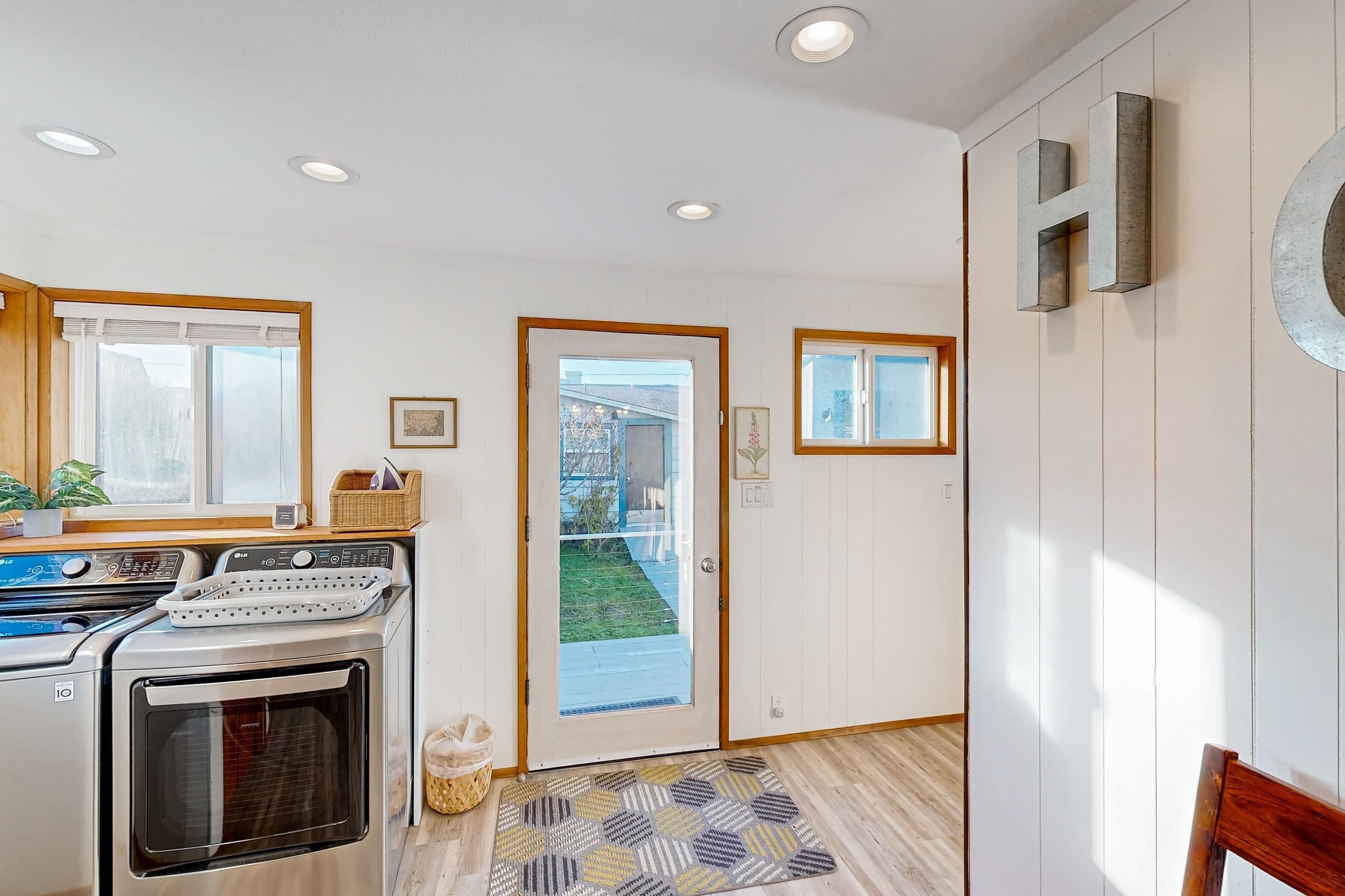A modern kitchen with a built-in oven and a washing machine, a wooden door leading to a garden, and a patterned rug on the floor.