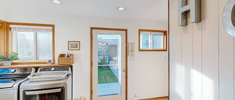 A modern kitchen with a built-in oven and a washing machine, a wooden door leading to a garden, and a patterned rug on the floor.