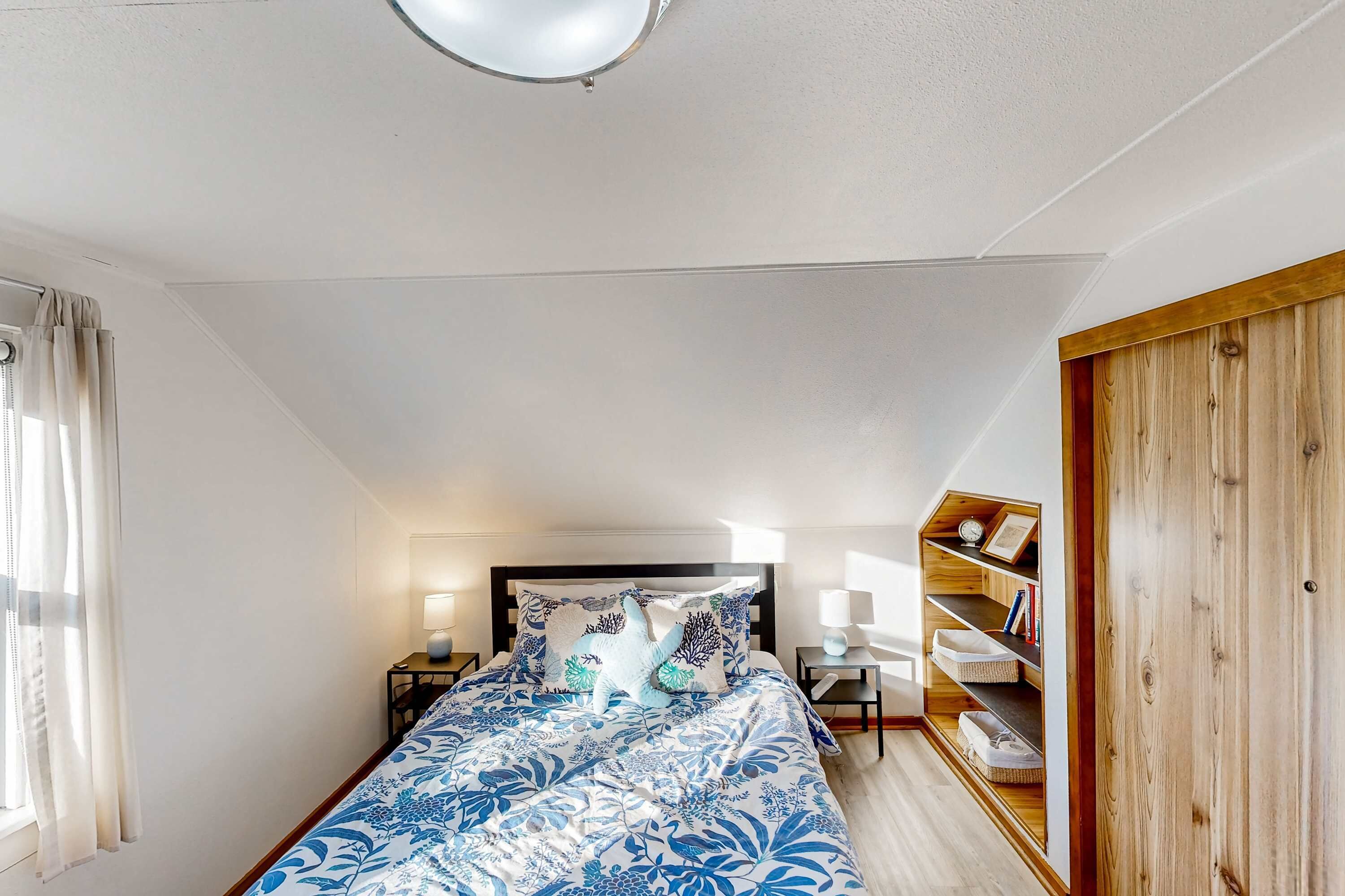 A bedroom with a bed, bedside tables, a nightstand, a wooden wardrobe, and a shelf with decorative items.