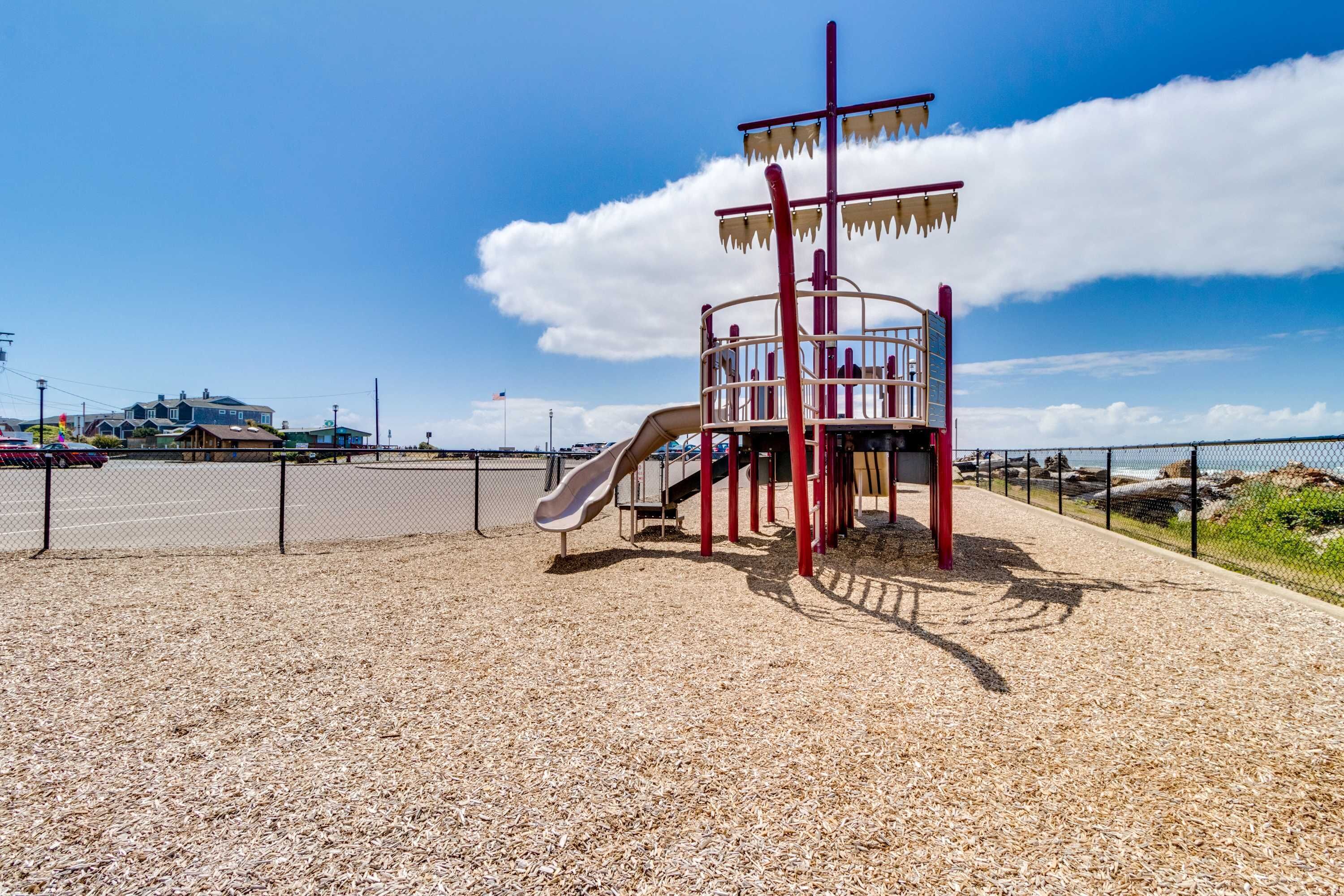 Un parque infantil con tobogán y estructura de escalada, rodeado por una cerca y una zona de grava.