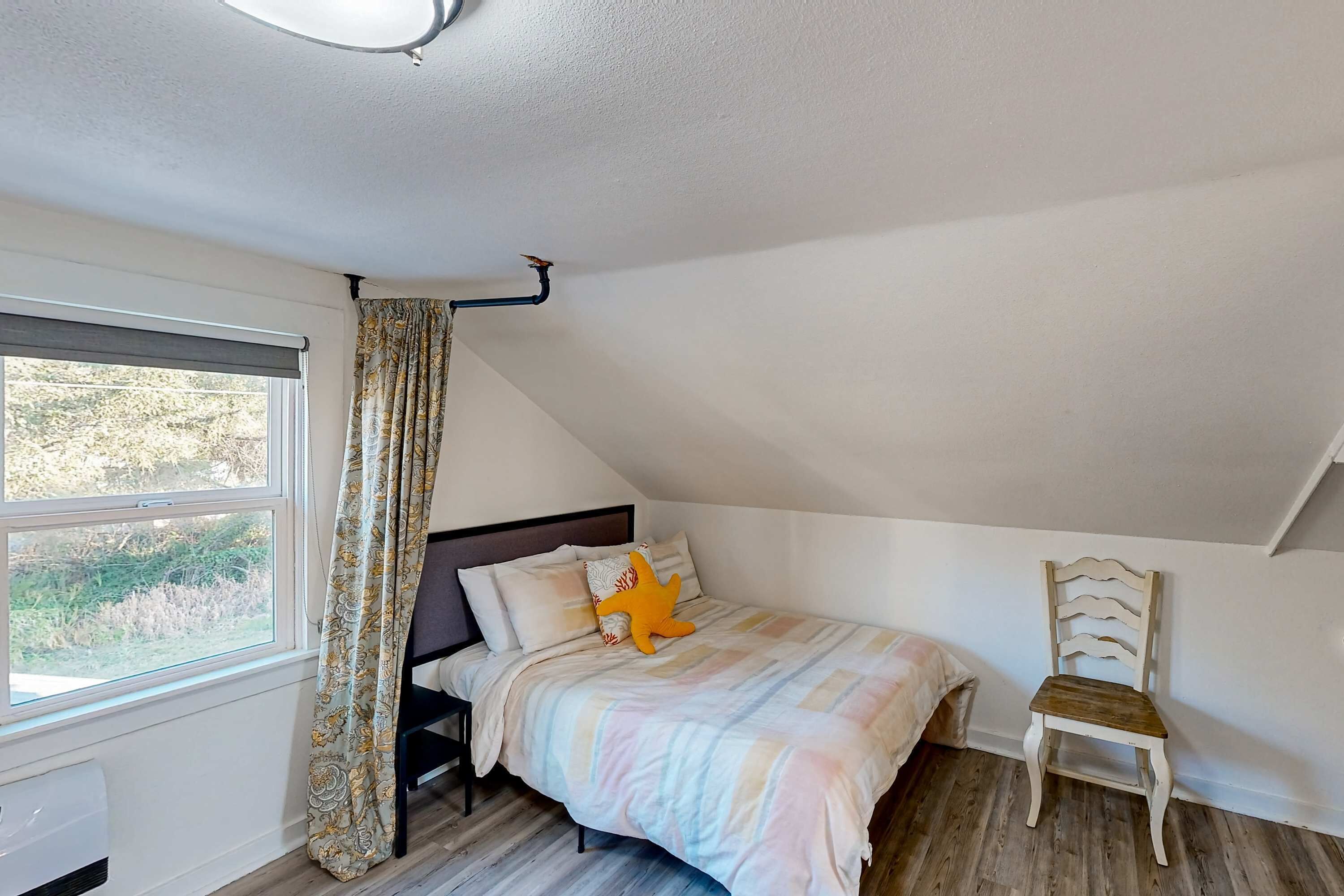 A bedroom with a sloped ceiling, a bed with a striped bedspread, a window with curtains, and a chair.