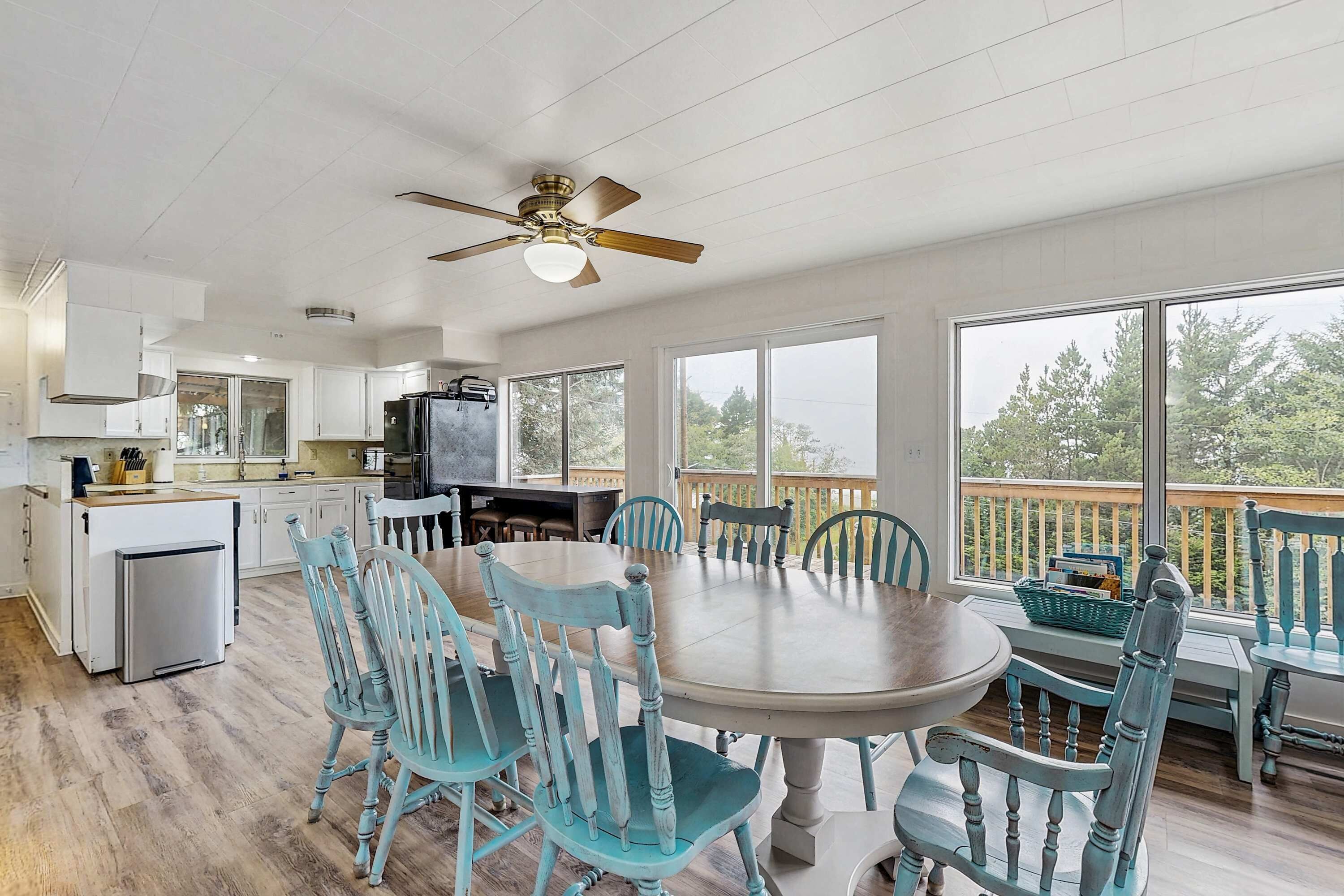A spacious kitchen with white cabinets, a dining area with a large table and blue chairs, and a balcony with a view of trees.