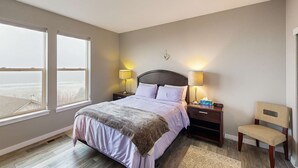 A bedroom with a bed, two bedside lamps, a nightstand, a chair, and a view through large windows. - Spacious retreat with beautiful ocean views dog friendly with WiFi patio (Rockaway Beach)
