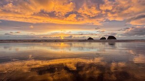 A beach with a sunset, calm waters, and three prominent rock formations in the distance.