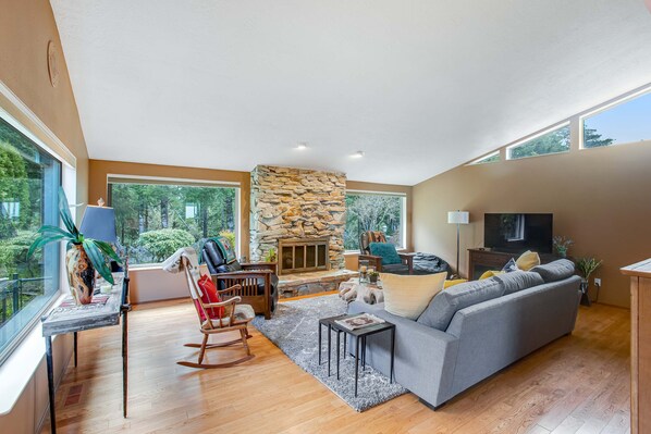 A spacious living room with a stone fireplace, large windows, a grey sofa, and a wooden floor. - Exquisite Home with Top of the World Views Free WiFi Washer Dryer (Tillamook)