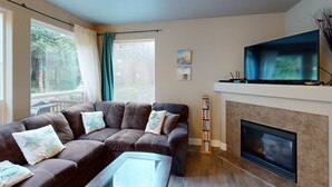 A living room with a brown sofa, a glass coffee table, a fireplace, and a flat-screen TV mounted above it. - Modern Ocean Highlands home with Netarts Bay and ocean views fireplace dogs OK (Tillamook)