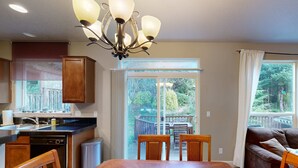 A dining area with a wooden table and chairs, a kitchen with a black countertop and wooden cabinets, and a view of a deck and greenery through a glass door. - Modern Ocean Highlands home with Netarts Bay and ocean views fireplace dogs OK (Tillamook)