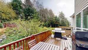 A wooden deck with a grill and a table, surrounded by a wooden fence and dense greenery. - Modern Ocean Highlands home with Netarts Bay and ocean views fireplace dogs OK (Tillamook)