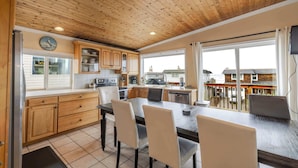 A kitchen with wooden cabinets, a dining table with chairs, and a view of the outside through large windows. - Ocean View Home with Rec Room Rock Wall and Private Hot Tub A Block From Beach (Rockaway Beach)