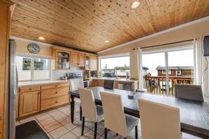 A kitchen with wooden cabinets, a dining table with chairs, and a view of the outside through large windows. - Ocean View Home with Rec Room Rock Wall and Private Hot Tub A Block From Beach (Rockaway Beach)