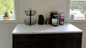 A kitchen with a white countertop, a dark wooden island, a coffee maker, a kettle, a jar, and a hanging wire basket.