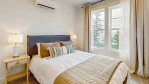 Una cama bien hecha con un edredón acolchado, dos lámparas de noche, una mesita y una ventana con cortinas.