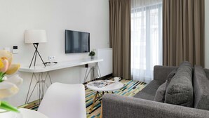 Una habitación de hotel moderna con un sofá gris, una mesa blanca, un televisor de pantalla plana y un amplio ventanal con cortinas.