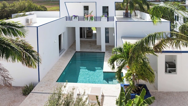 A modern white building with a swimming pool, surrounded by palm trees and a clear sky. - Villa Santorini – Luxury 4-Bedroom Villa with Pool, Gym & Outdoor Living in Brakkeput, Curaçao (Willemstad)