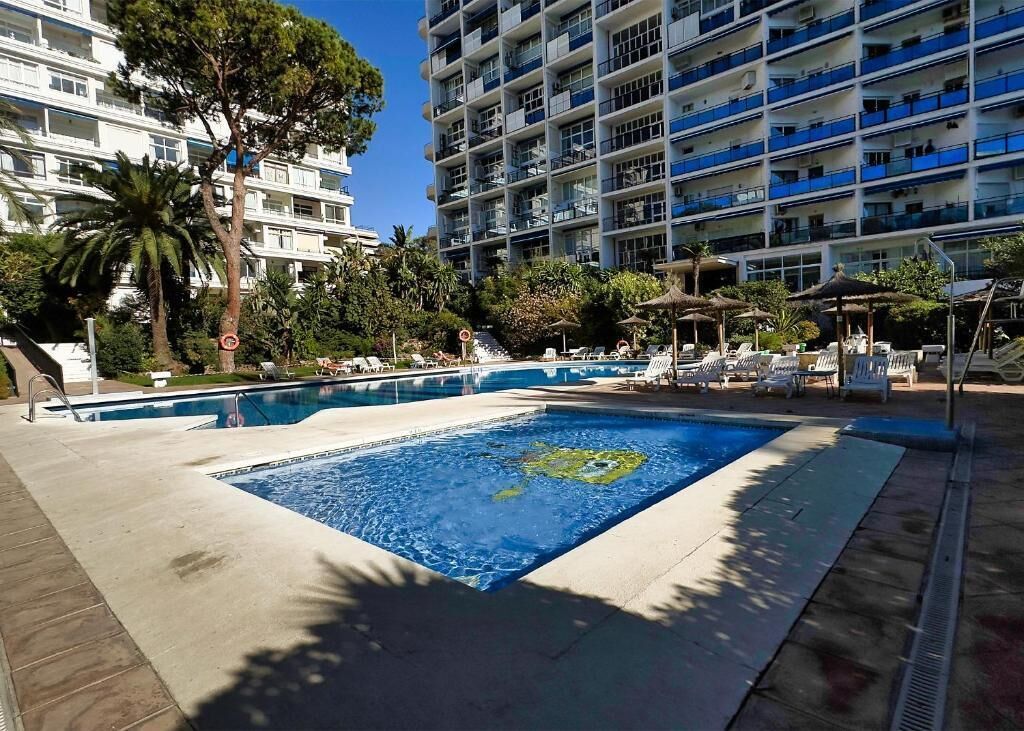 A swimming pool with lounge chairs and umbrellas, surrounded by tall buildings and palm trees.