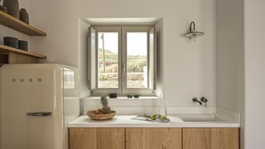A kitchen with a white refrigerator, wooden cabinets, a window with a view, and a wall-mounted light fixture. - Casa Botanica · Heather (Antiparos)