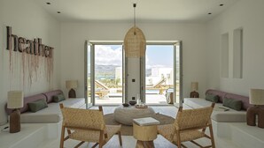 A spacious living room with a large window, two wicker chairs, a small wooden table, and a sofa with cushions. - Casa Botanica · Heather (Antiparos)