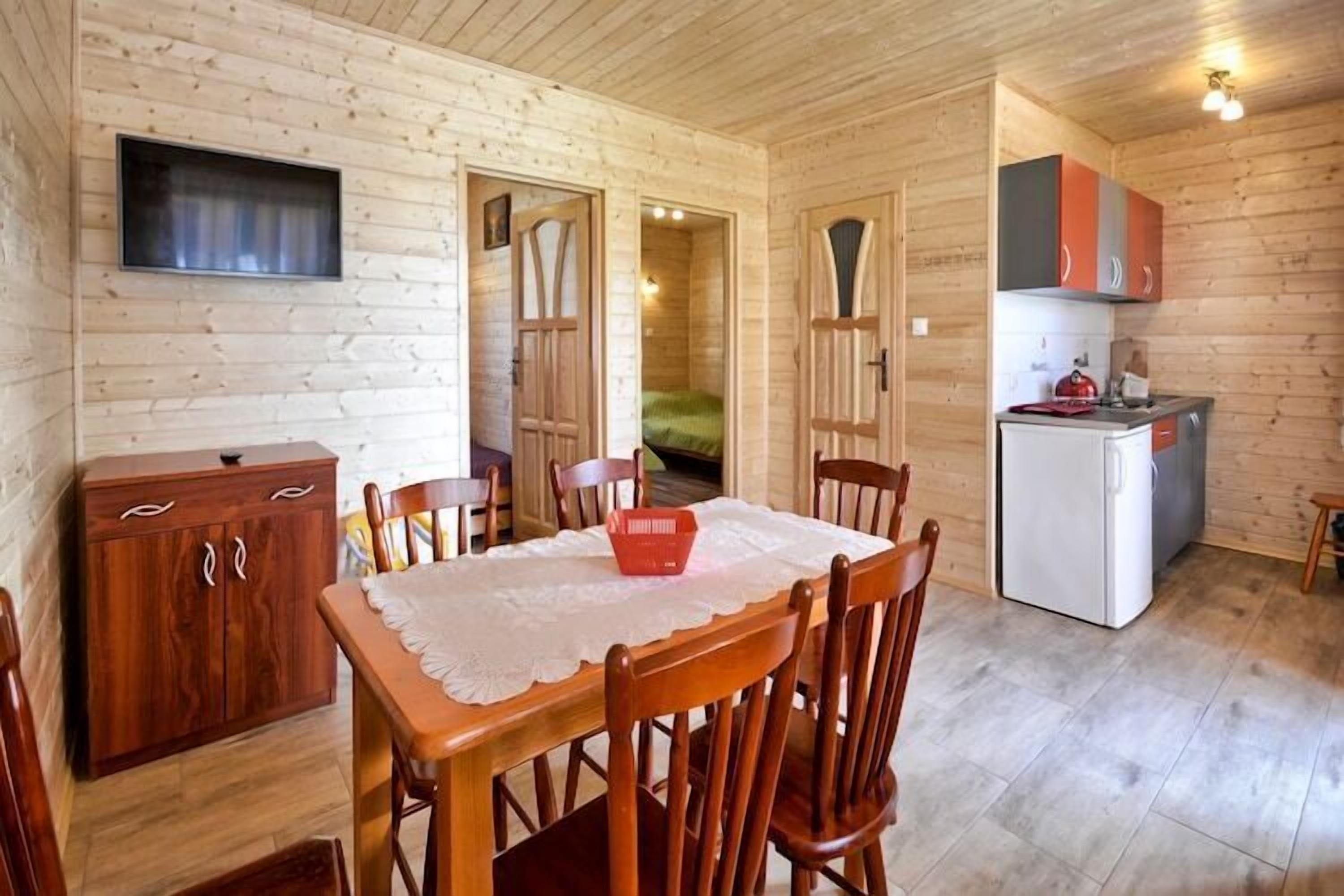 A wooden cabin interior with a dining table, chairs, a kitchen area with cabinets, a refrigerator, and a television mounted on the wall.