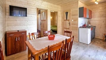 A wooden cabin interior with a dining table, chairs, a kitchen area with cabinets, a refrigerator, and a television mounted on the wall.