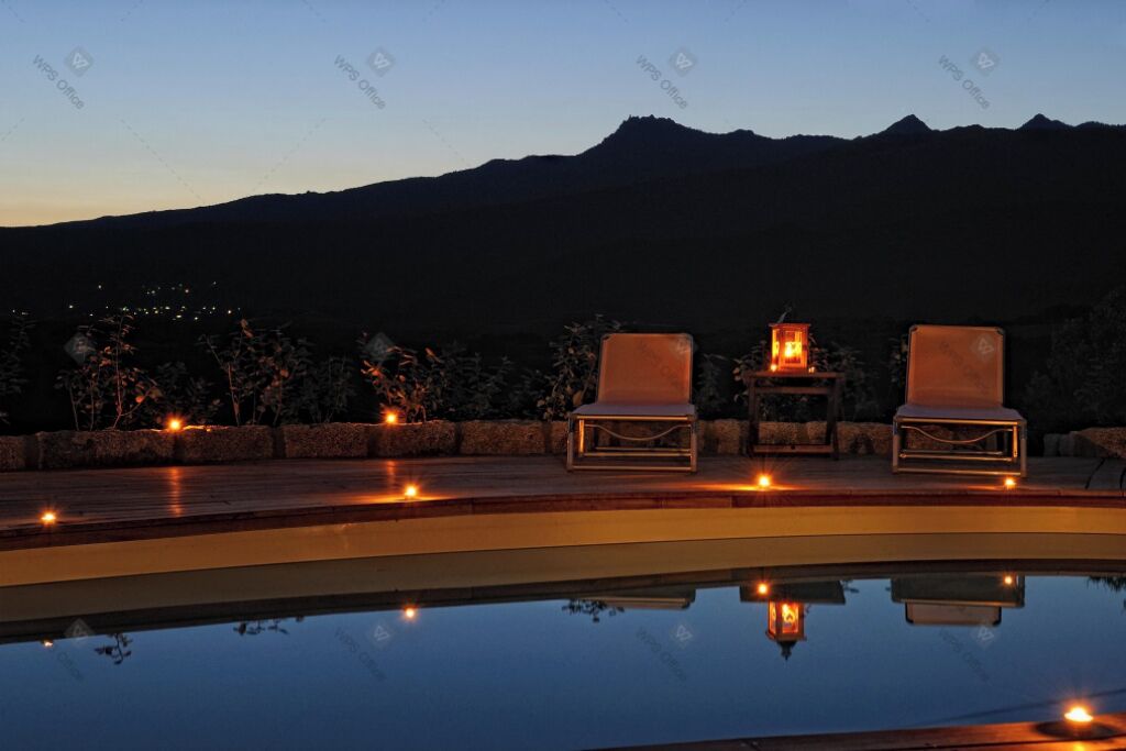 A poolside area with two lounge chairs and a table, surrounded by a low wall and a mountain backdrop.