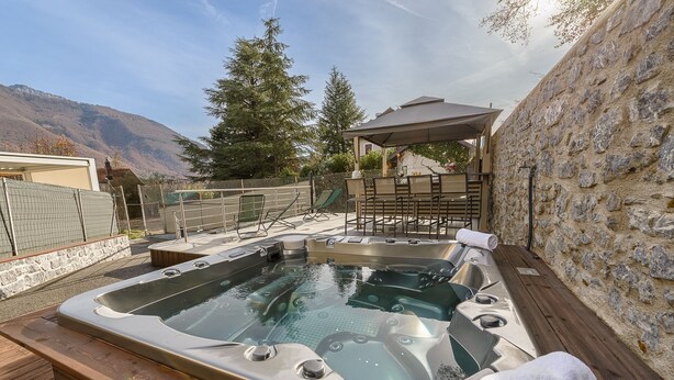 A hot tub area with a wooden deck, stone wall, and a view of mountains. - La Source du Lac - Plage privée (Doussard)