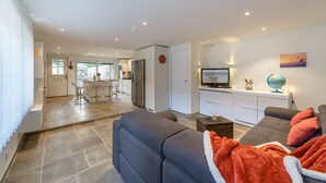 A modern living room with a grey sofa, a coffee table, and a TV. There is a kitchen in the background with a fridge, stove, and bar stools. - La Source du Lac - Plage privée (Doussard)