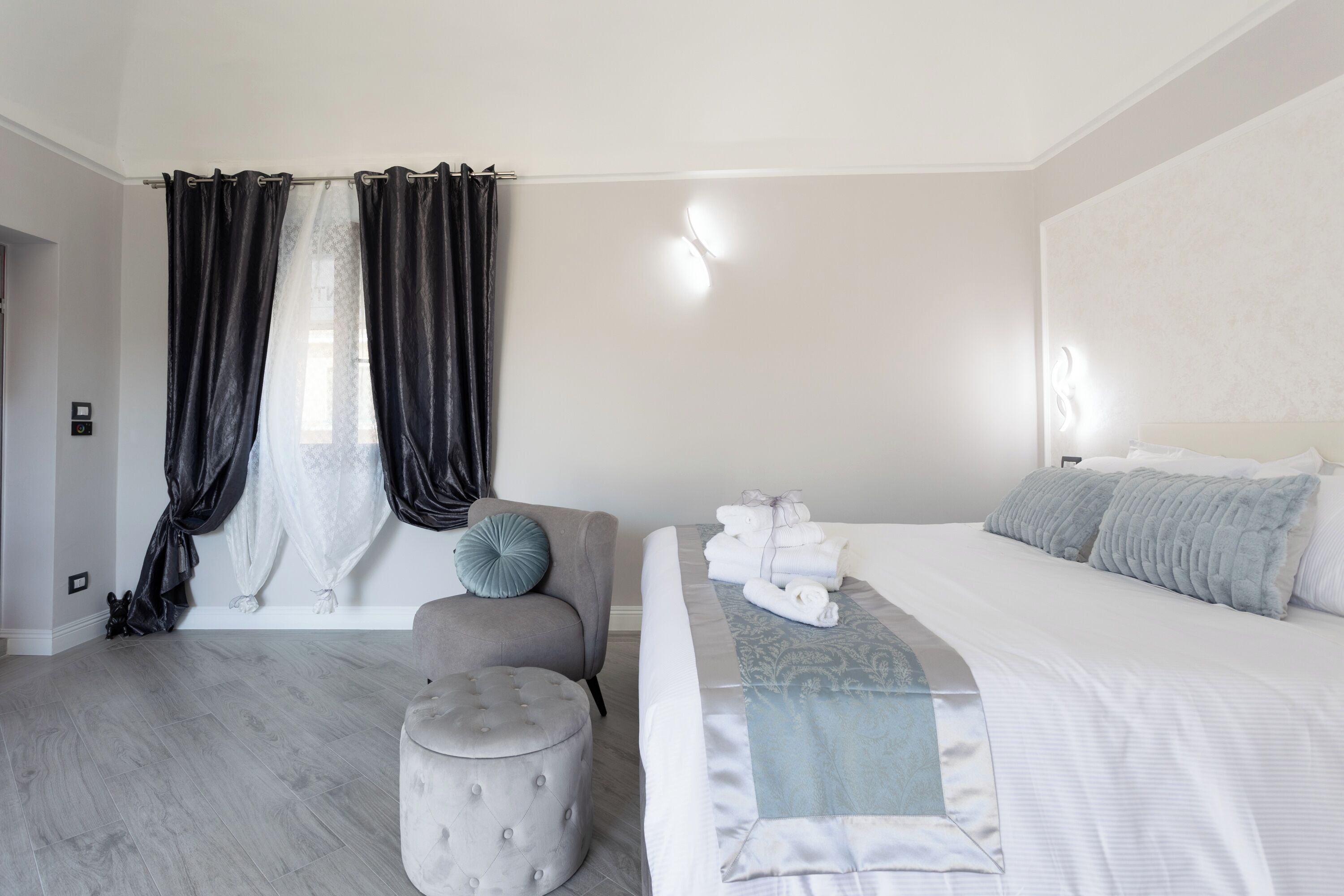 Ein modernes Schlafzimmer mit einem großen Bett, einem grauen Sessel, einem kleinen Hocker und einem Fenster mit schwarzen Vorhängen.