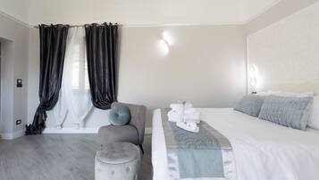 Ein modernes Schlafzimmer mit einem großen Bett, einem grauen Sessel, einem kleinen Hocker und einem Fenster mit schwarzen Vorhängen.